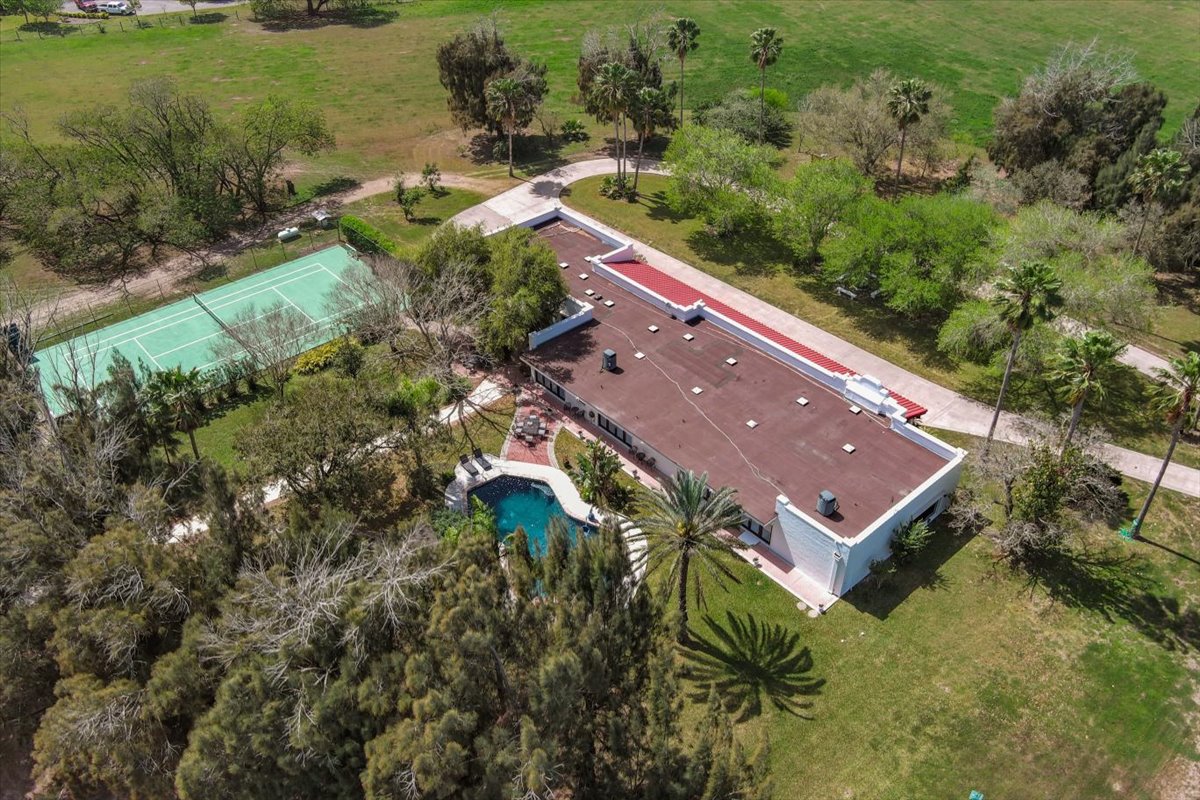 Aerial view of a single-story house with a flat roof, surrounded by trees, a swimming pool, and a tennis court in a grassy area.