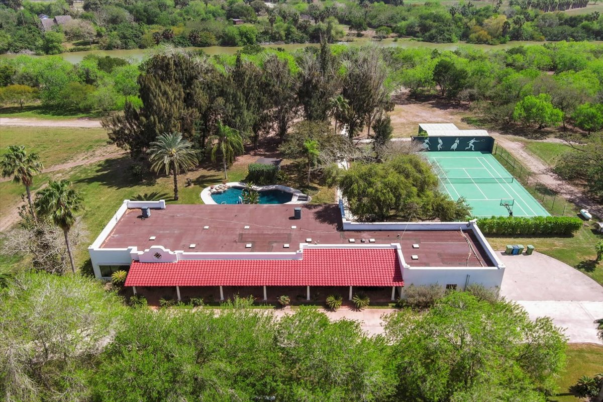 Aerial view of a single-story building with a red-tiled roof, a swimming pool, and a fenced tennis court surrounded by trees and greenery.