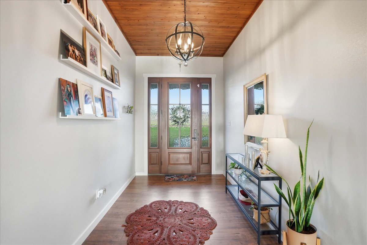 Entrance hallway with wooden front door, decorative round rug, photo shelves on left wall, and a console table with lamp and plants on right.