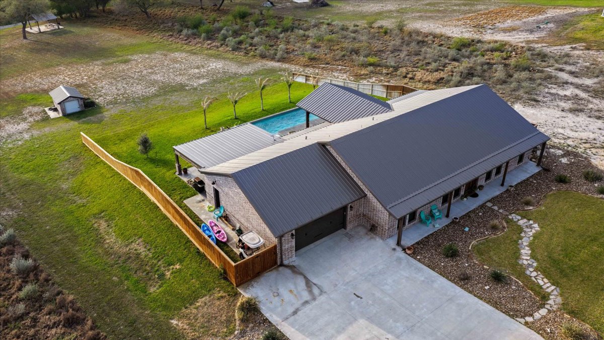 Aerial view of a single-story house with a gray metal roof, a fenced backyard with a swimming pool, and a concrete driveway.