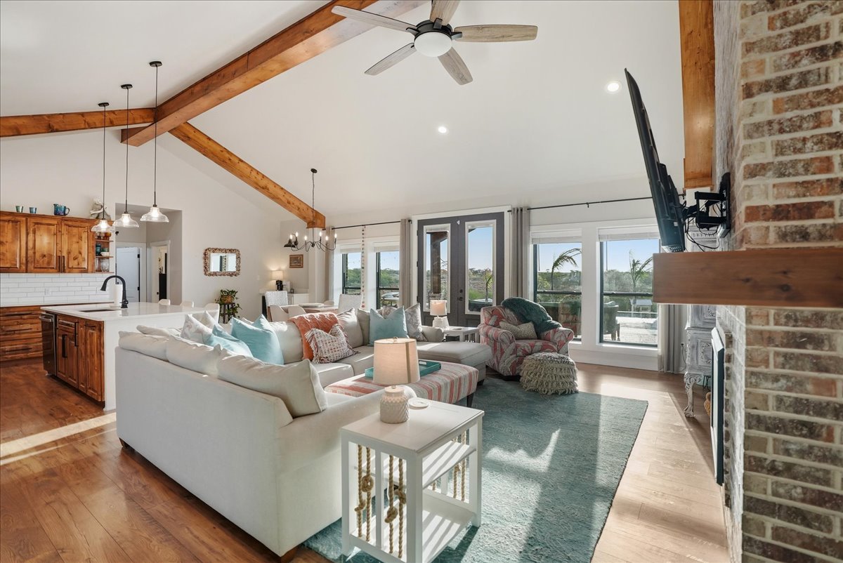 Bright living room with vaulted ceiling, wooden beams, white sectional sofa with colorful pillows, floral armchair, a ceiling fan, and large windows with a patio view.