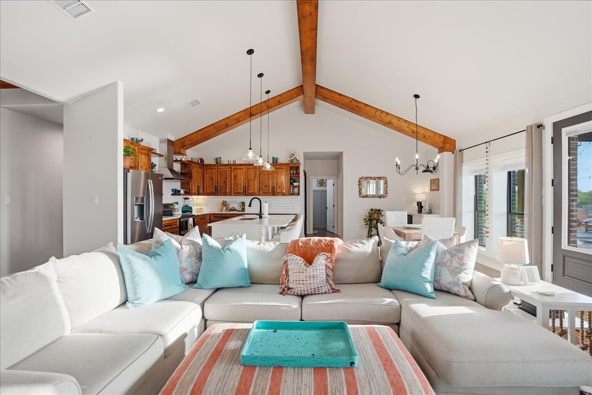 Bright open-concept living room and kitchen with vaulted ceiling, wooden beams, light beige sectional sofa with blue and patterned pillows, and wooden kitchen cabinets.