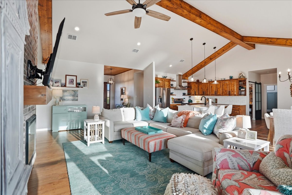 Bright open living room with large beige sectional sofa, blue area rug, colorful ottoman, wooden kitchen with island, and exposed ceiling beams.
