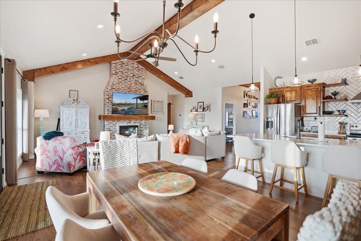 Open-concept living and kitchen space with wooden dining table, white chairs, exposed wood beams, brick fireplace, and a television mounted above.