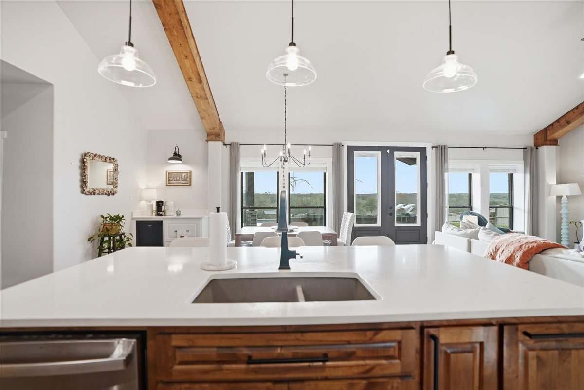 Modern open-concept living area with a white kitchen island sink, wooden lower cabinets, dining table, and large windows with grey curtains overlooking greenery.