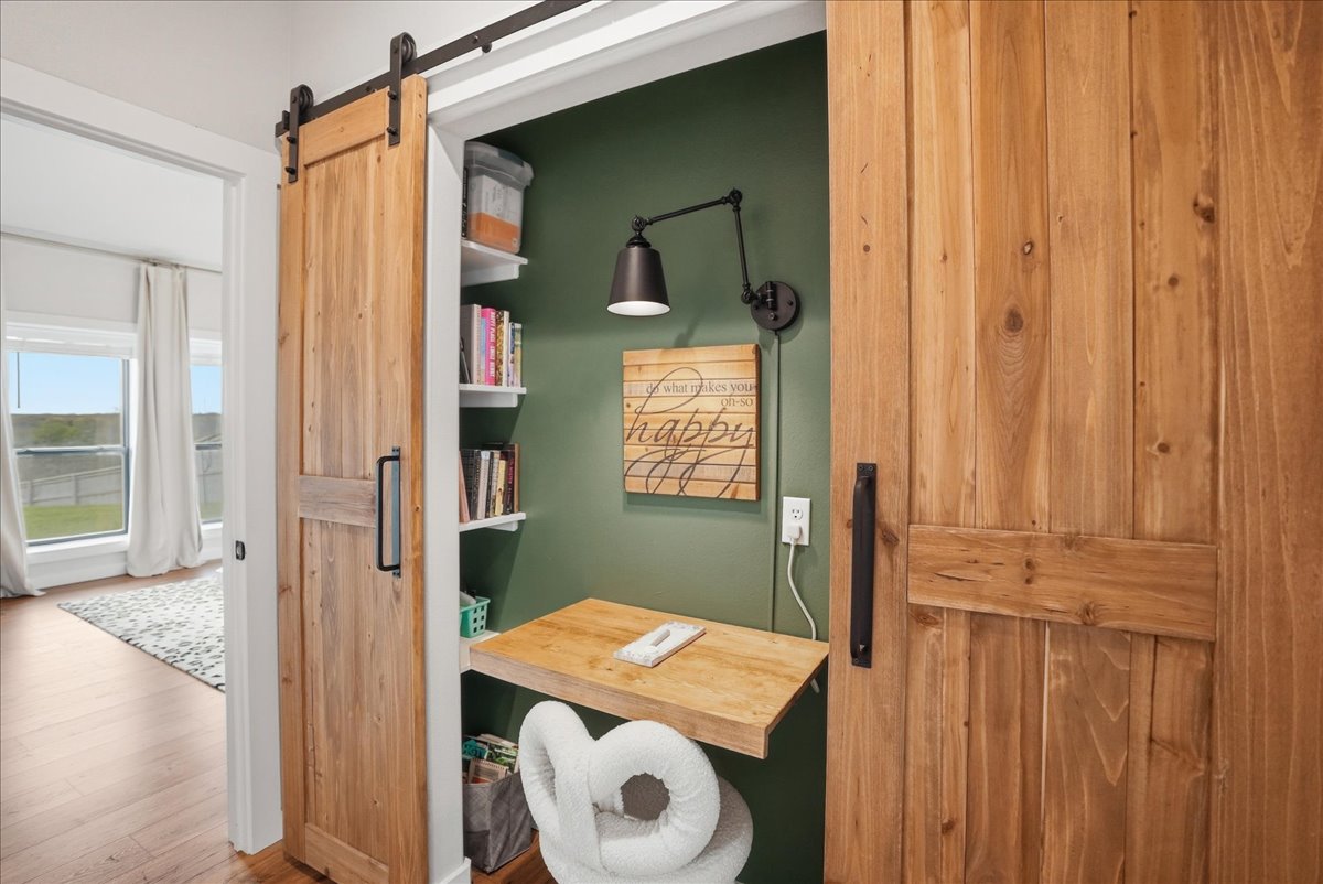 Small cozy workspace closet with green walls, wooden sliding barn doors, a floating wooden desk, a white cushioned chair, shelves with books, and a wall-mounted black lamp.