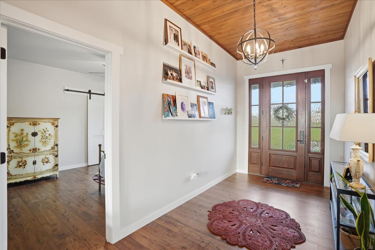 Entryway with wooden floor and ceiling, a round red rug, wall-mounted photo shelves, a chandelier, and wooden double front doors with a wreath.