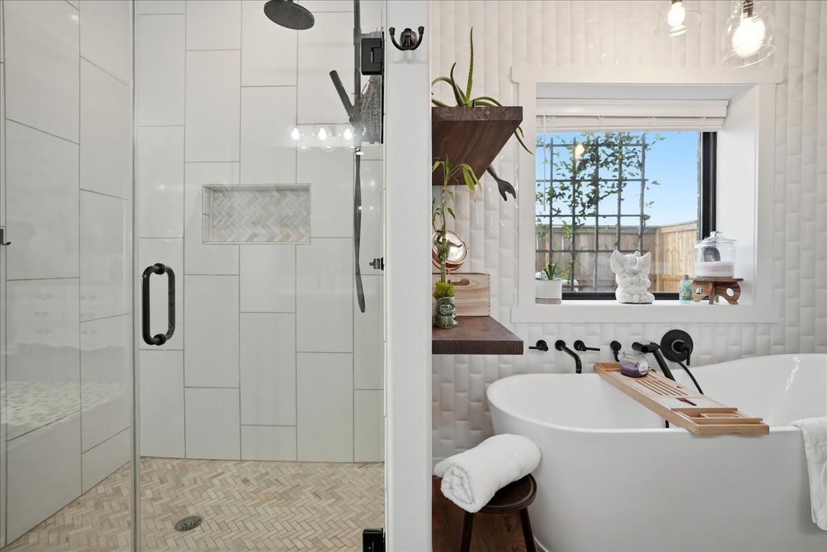 Modern bathroom with a glass-enclosed shower on the left and a white freestanding bathtub with wooden shelves and a window on the right.