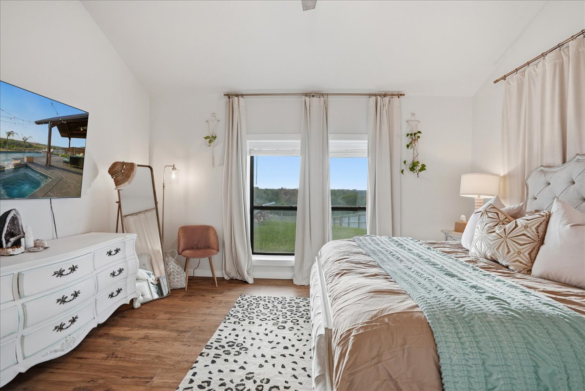 Bright bedroom with a large bed adorned with beige and light blue bedding, a white dresser, wall-mounted TV, leopard print rug, and two windows with white curtains overlooking greenery.