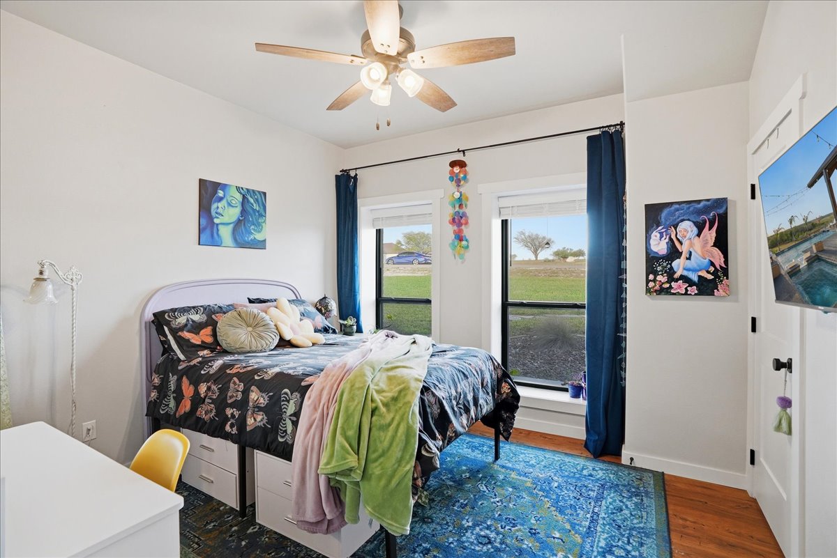 Cozy bedroom with a bed featuring butterfly-patterned bedding, green and pink blankets, blue curtains, two paintings on the walls, and large windows overlooking a green lawn.