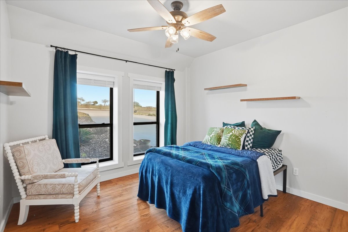 Bedroom with a double bed dressed in blue and green bedding, decorative pillows, two windows with dark blue curtains, wooden floors, a cream armchair, and ceiling fan with wooden blades.