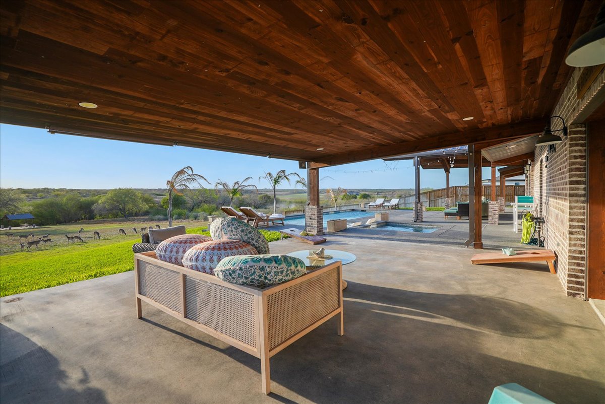 Covered patio with cushioned outdoor sofa overlooking a grassy field with deer and a swimming pool in the background.