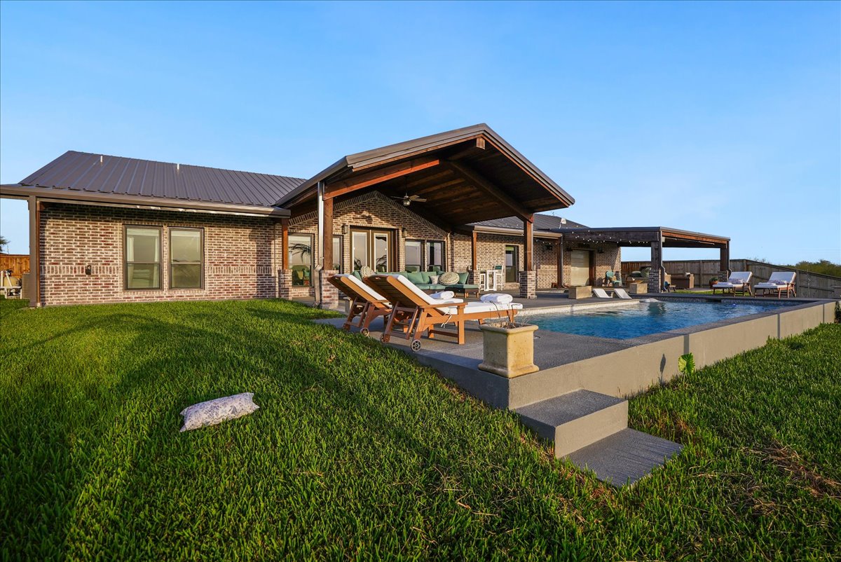 Modern brick house with a covered patio, outdoor lounge chairs, and a rectangular swimming pool surrounded by green grass.