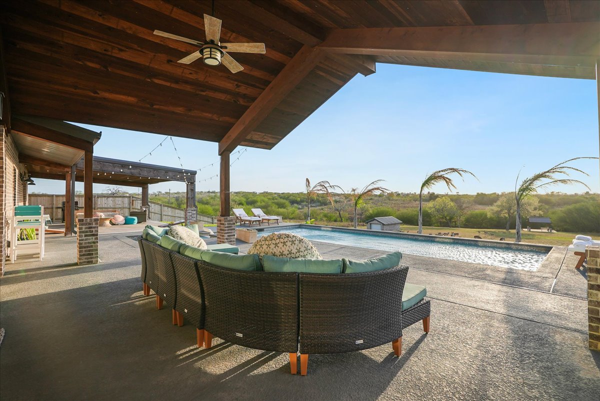 Covered patio with ceiling fan, wicker sectional sofa with green cushions, overlooking an outdoor swimming pool and open green landscape.