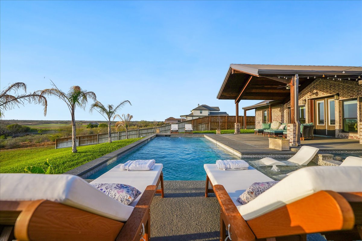Outdoor swimming pool with lounge chairs and towels, next to a covered patio attached to a brick house, under a clear blue sky.