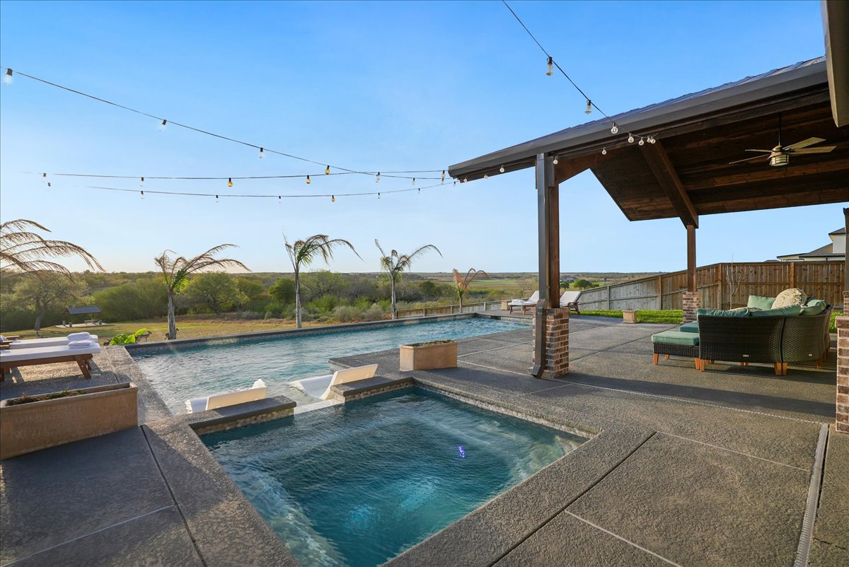 Outdoor pool area with an attached spa, surrounded by a concrete deck, lounge chairs, and a covered patio with seating under string lights and a clear blue sky.