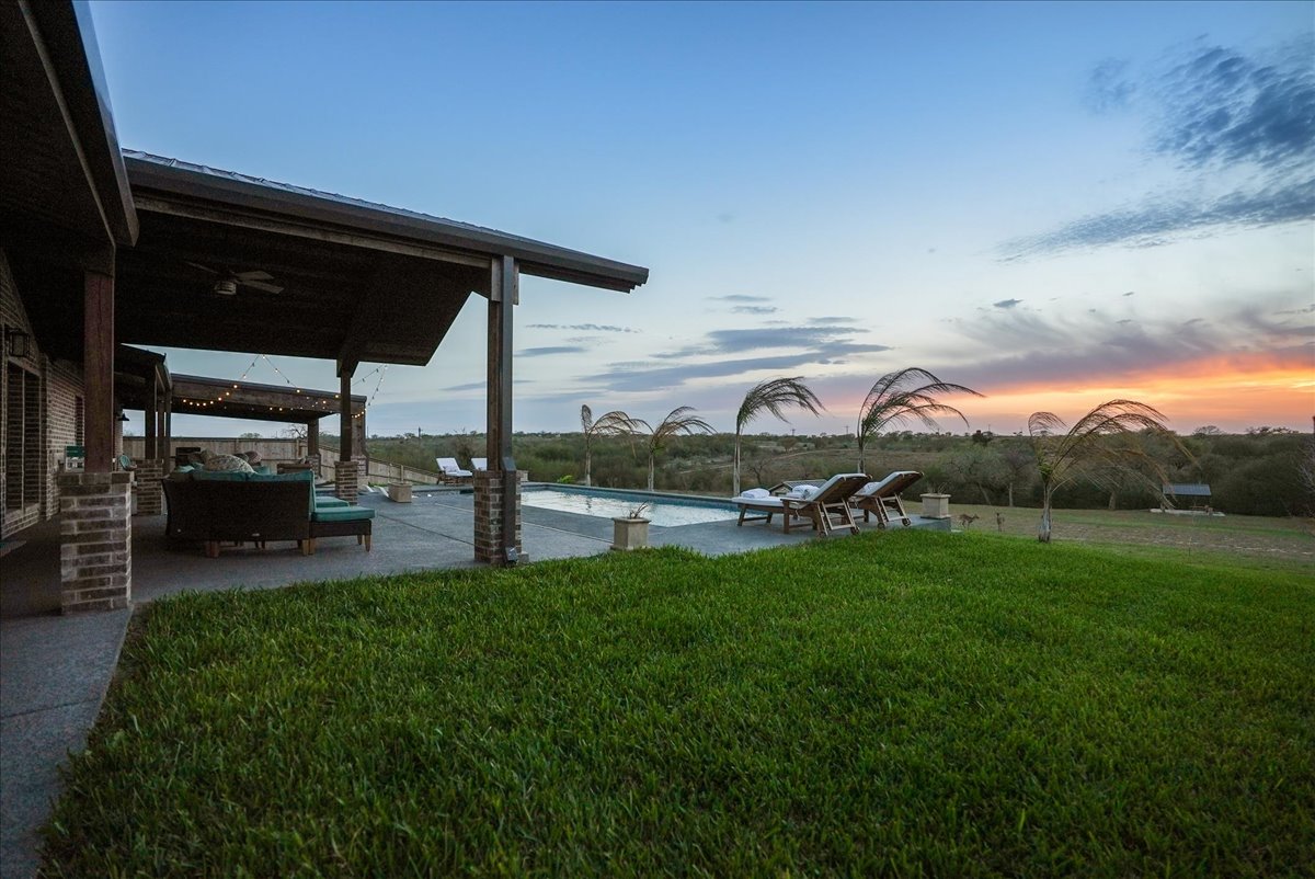 Backyard with covered patio, seating area, swimming pool, lounge chairs, and grassy yard at sunset.