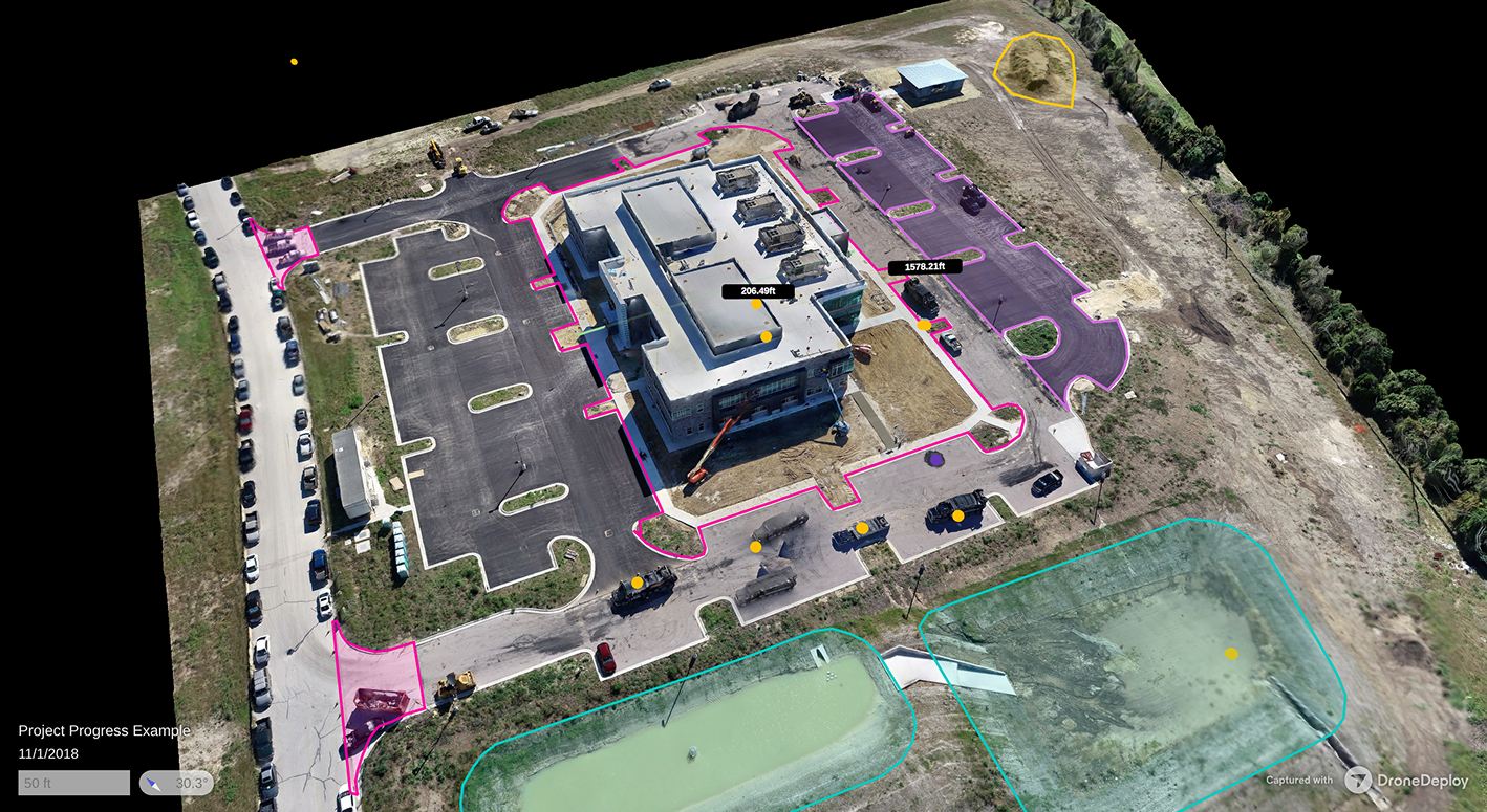 Aerial view of a construction site showing a large building with surrounding parking lots, marked measurements, vehicles, dirt piles, and water retention ponds outlined in different colors.