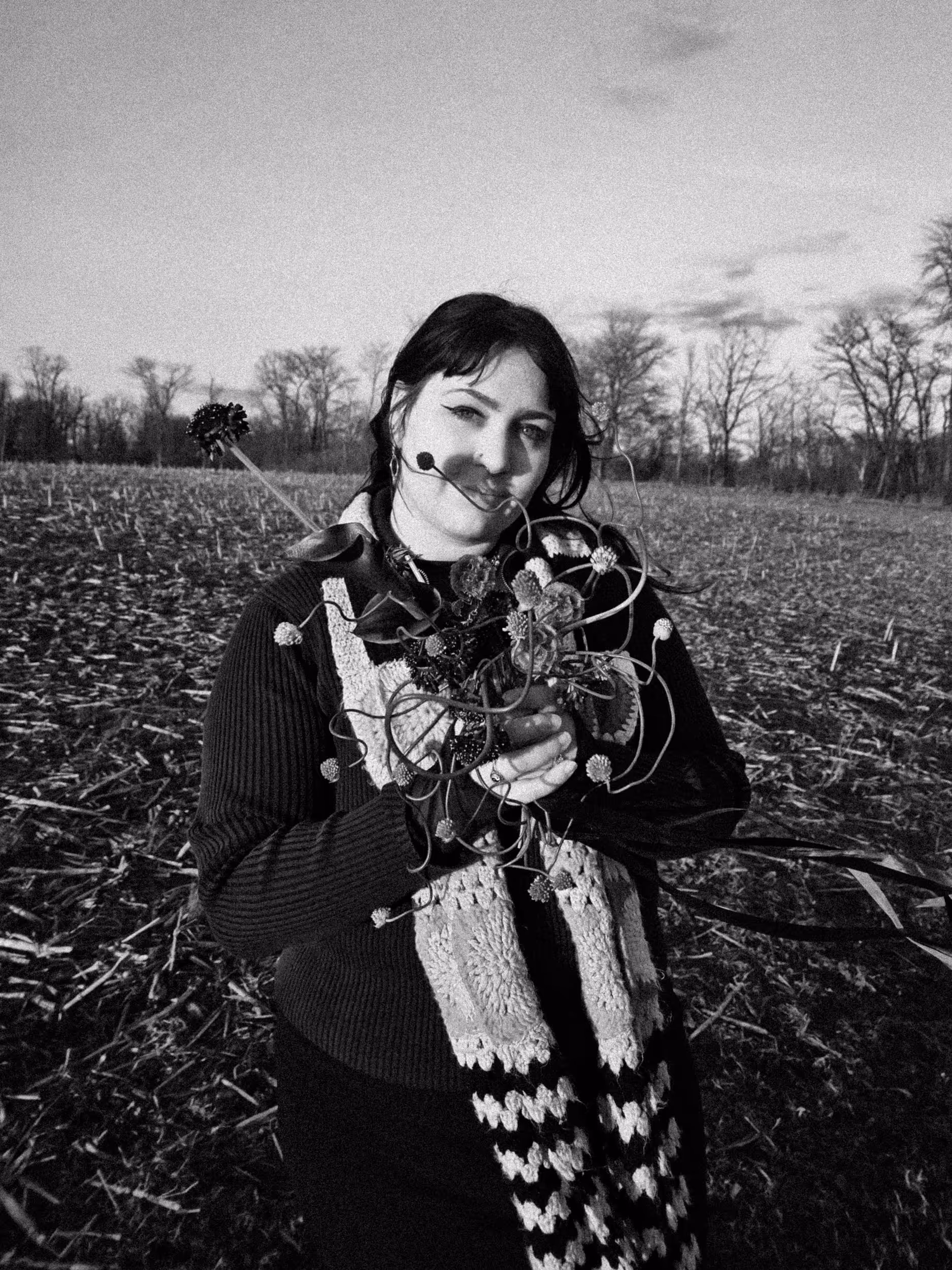 Young woman in a field holding a bouquet of wildflowers and vines, wearing a knitted scarf and sweater.