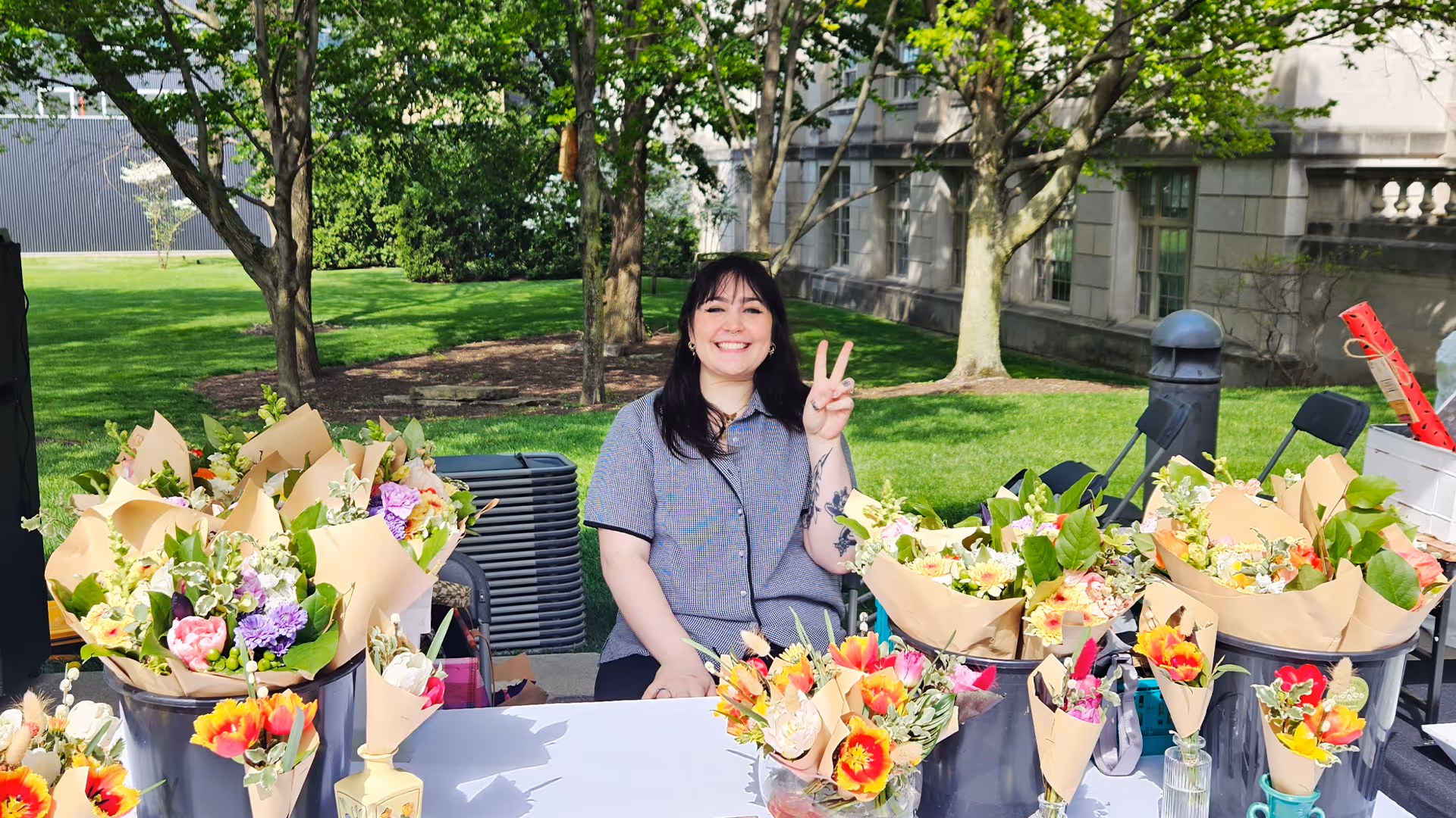 Smiling woman sitting behind a table with multiple bouquets of colorful flowers wrapped in brown paper outdoors.