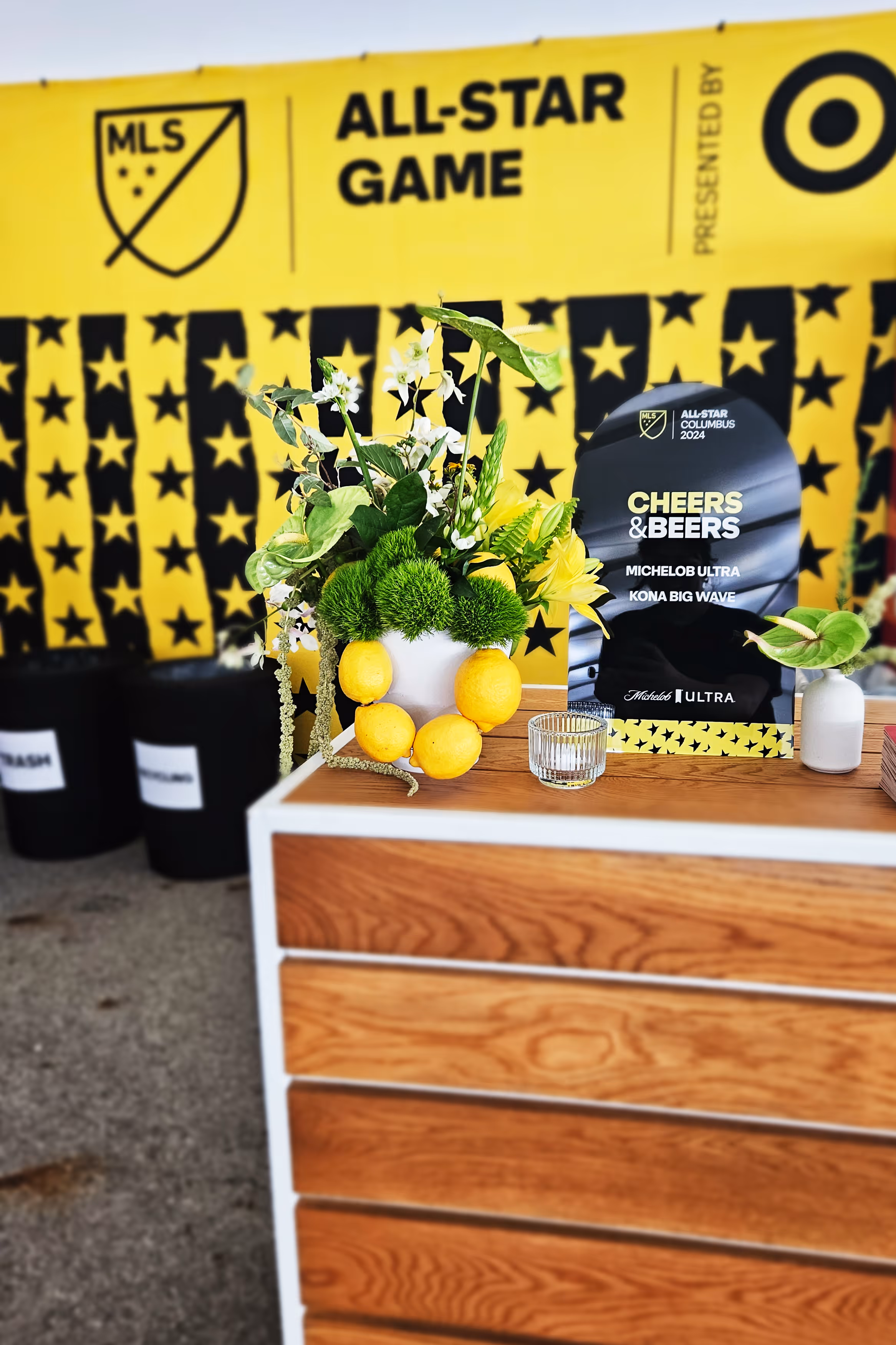 Decorative flower and lemon arrangement on a wooden counter with a black sign promoting the 2024 MLS All-Star Game and beers.