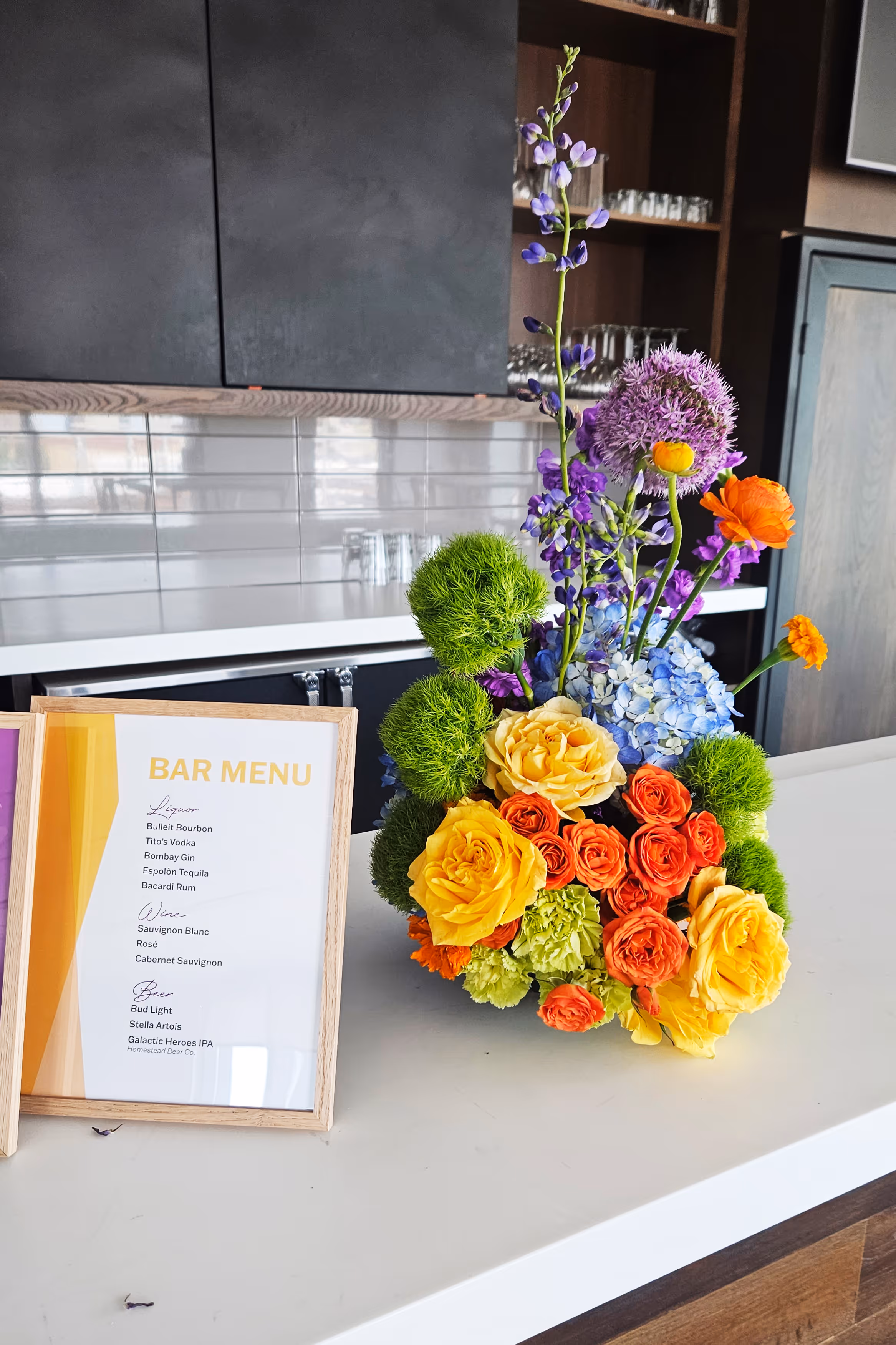 Colorful floral arrangement with yellow, orange, green, purple, and blue flowers next to a framed bar menu on a white countertop in a kitchen.
