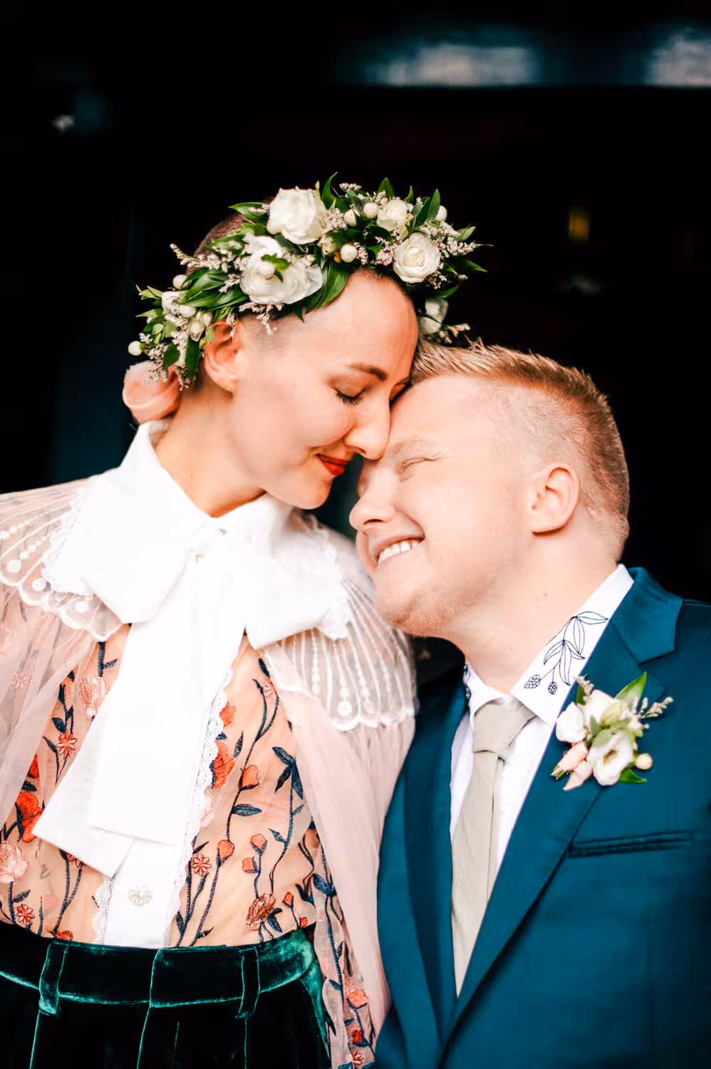 Couple with foreheads touching, the woman wearing a flower crown and embroidered blouse, and the man in a teal suit with floral collar details and boutonniere.