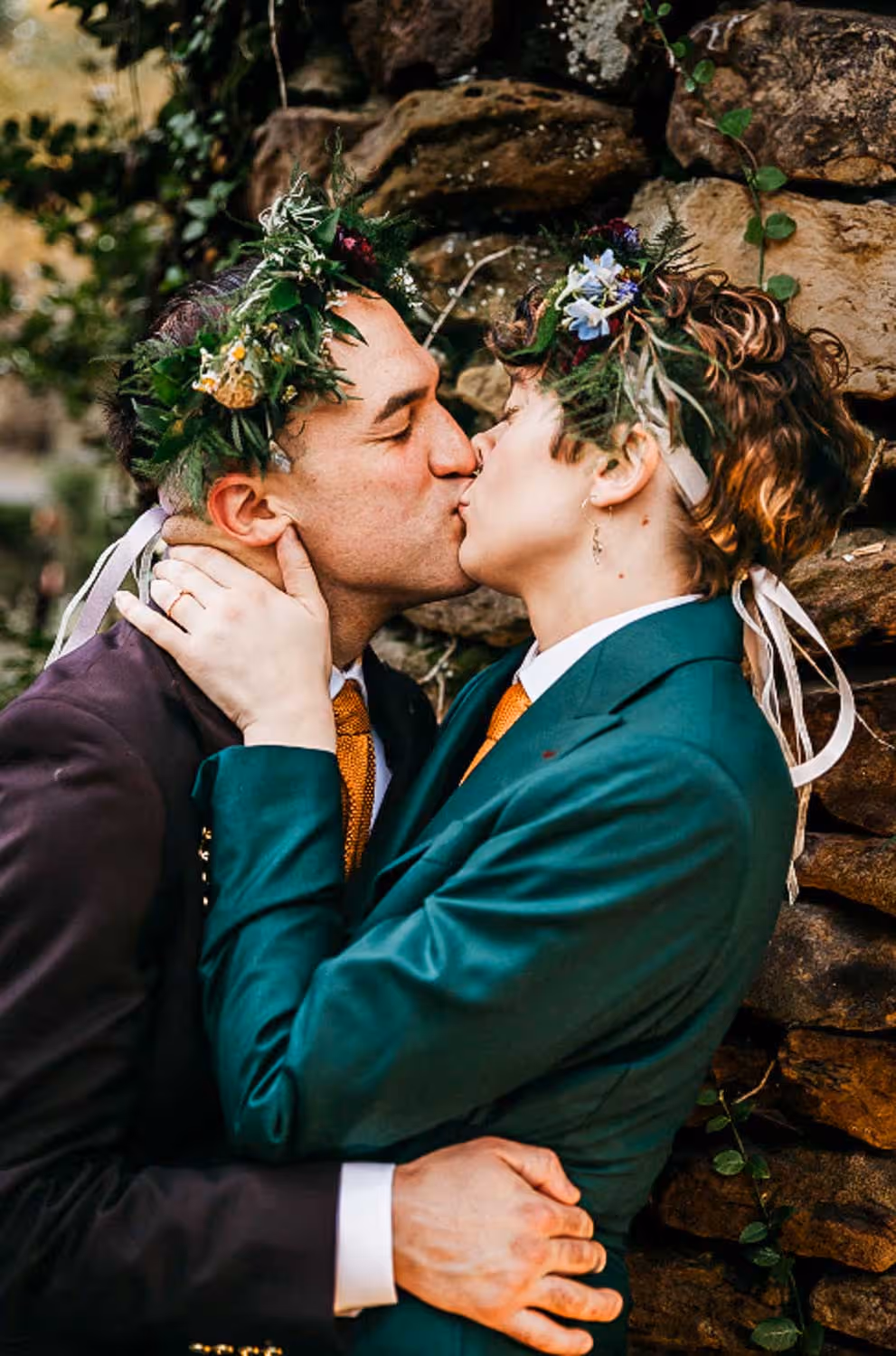 Couple wearing floral crowns and formal suits kissing in front of a stone wall.