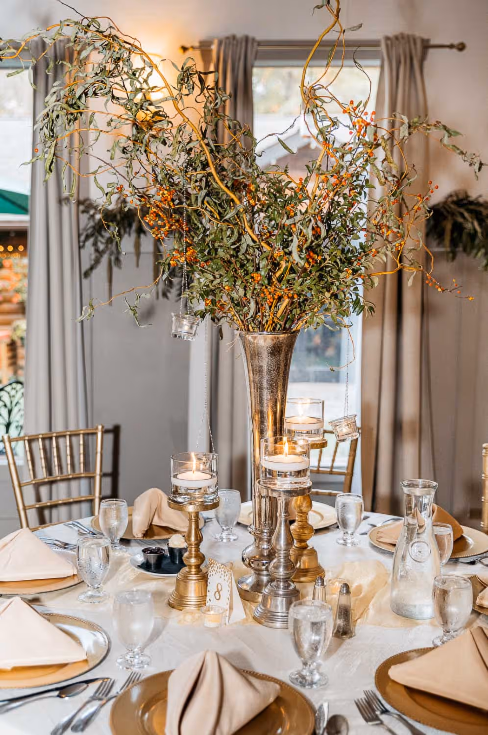 Elegant round dining table set with beige napkins on gold plates, glassware, and a tall silver vase centerpiece with green branches and orange berries, surrounded by lit candles on gold and silver holders.