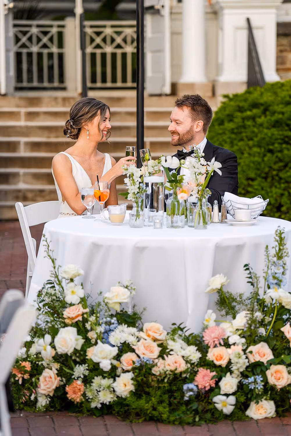 Bride and groom smiling and clinking champagne glasses at a decorated wedding reception table with white tablecloth and floral arrangements.