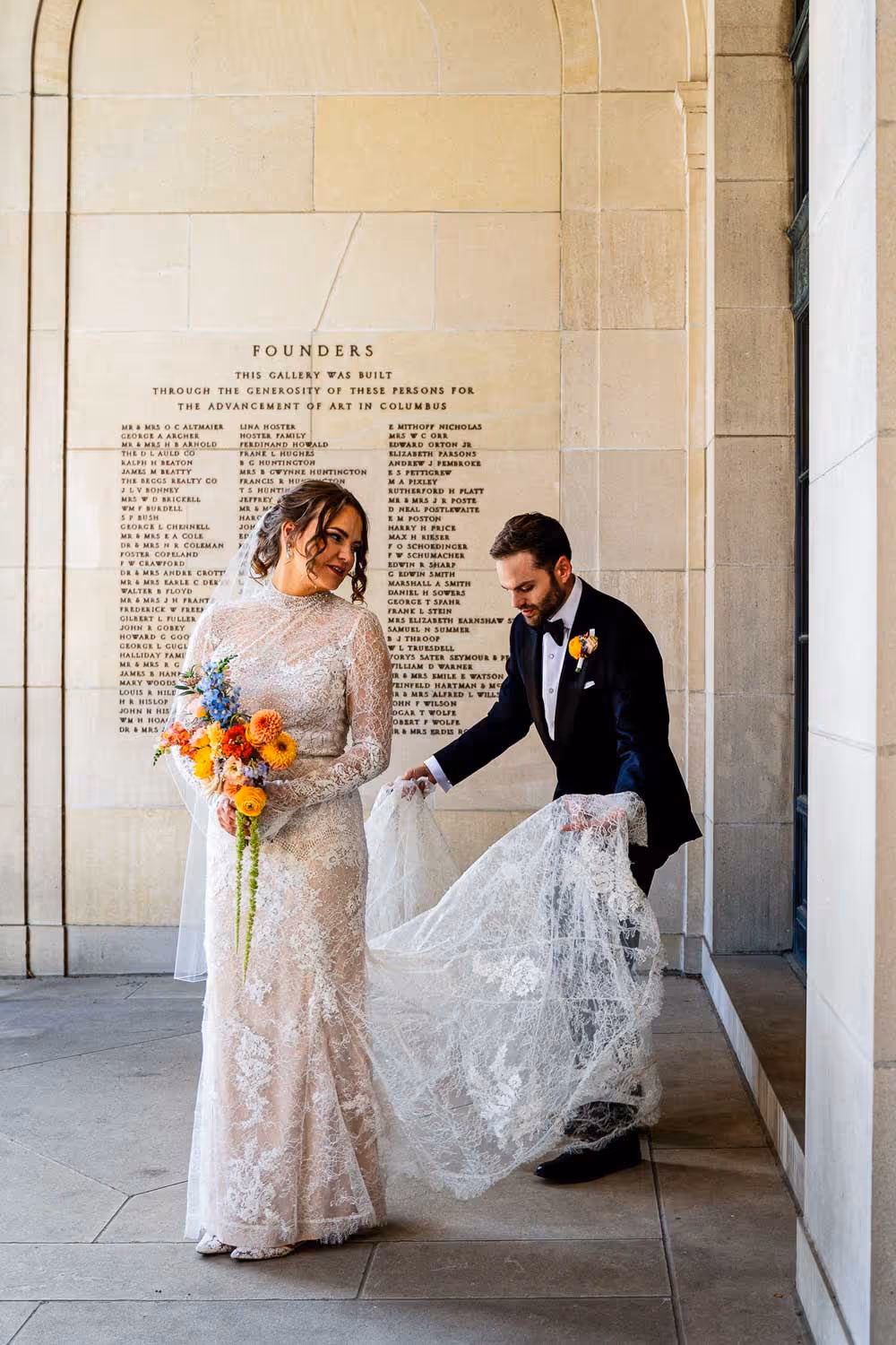 Bride in a lace wedding dress holding a vibrant bouquet, with groom in a black tuxedo adjusting her dress train in a stone gallery.