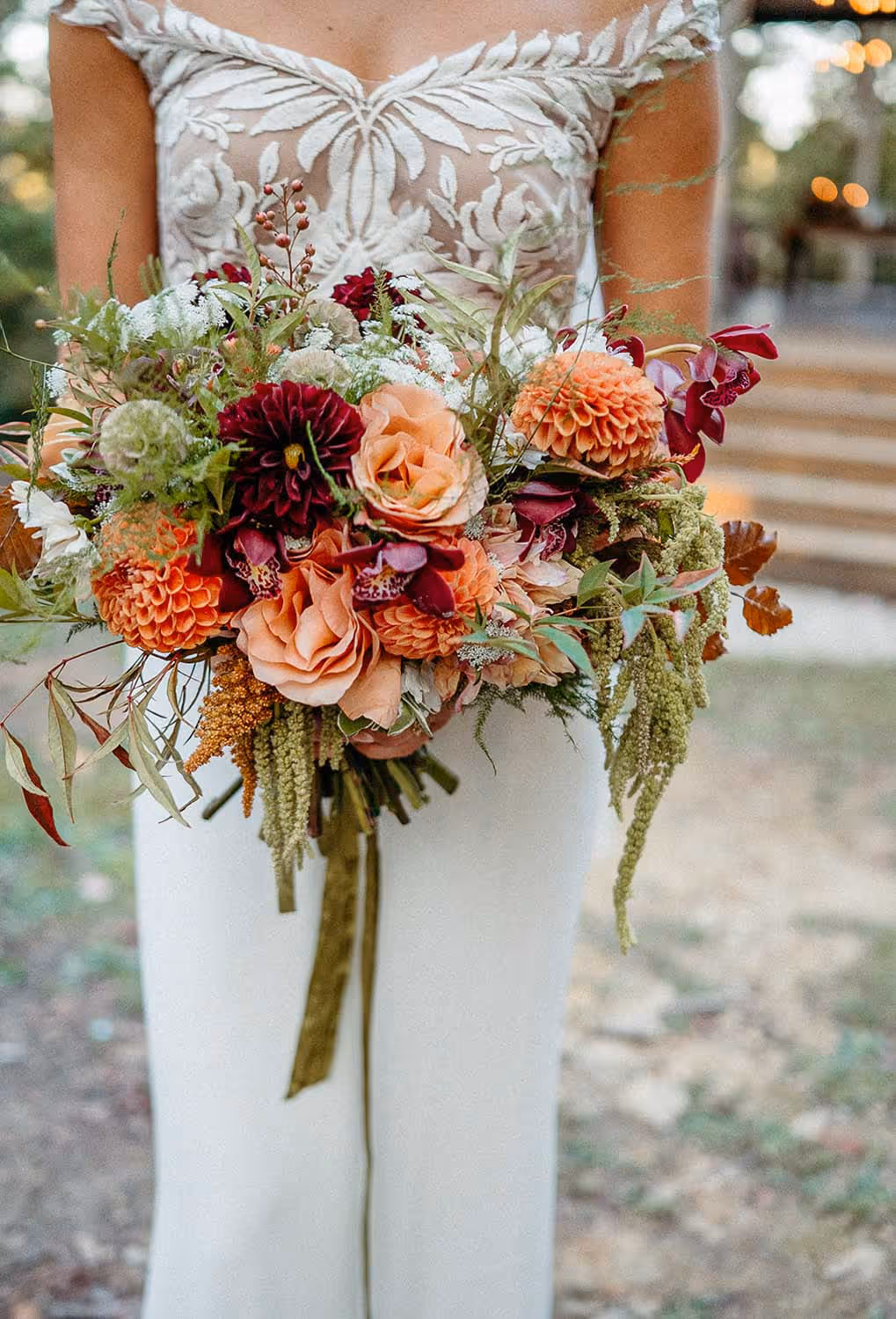 Bride in white gown holding a bouquet of mixed flowers including peach roses, burgundy and orange blooms, and green foliage.