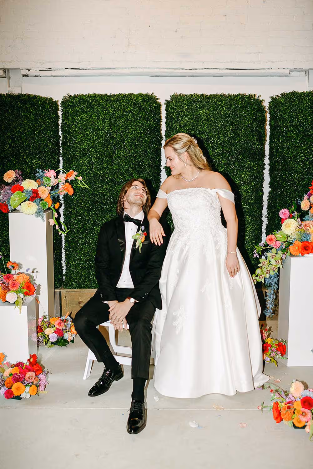 Bride in a white off-shoulder wedding gown standing next to groom in a black tuxedo sitting on a white chair, surrounded by colorful floral arrangements and green hedge panels.