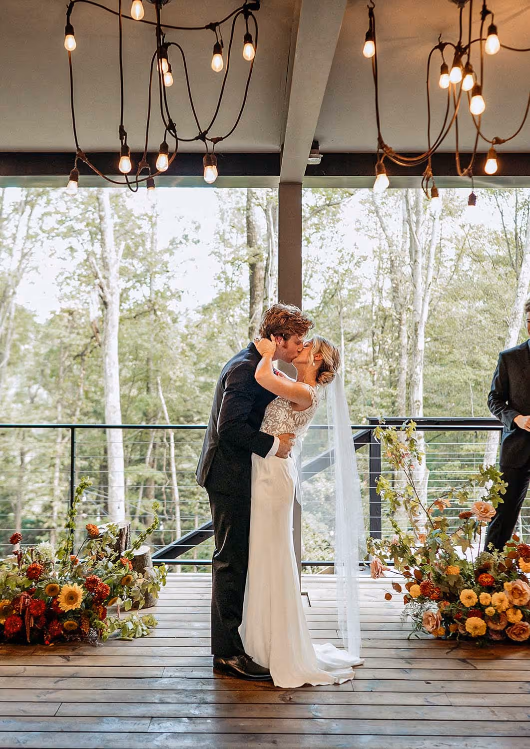 Bride and groom kissing during their wedding ceremony on a wooden deck decorated with floral arrangements and hanging lights.