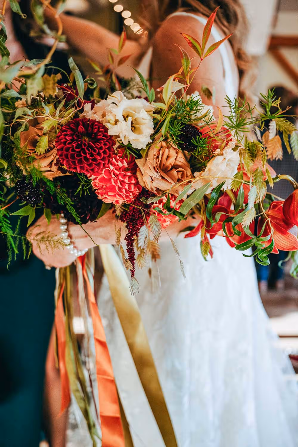 Close-up of a bride holding a vibrant wedding bouquet with red, cream, and peach flowers accented by green foliage and ribbon strands.