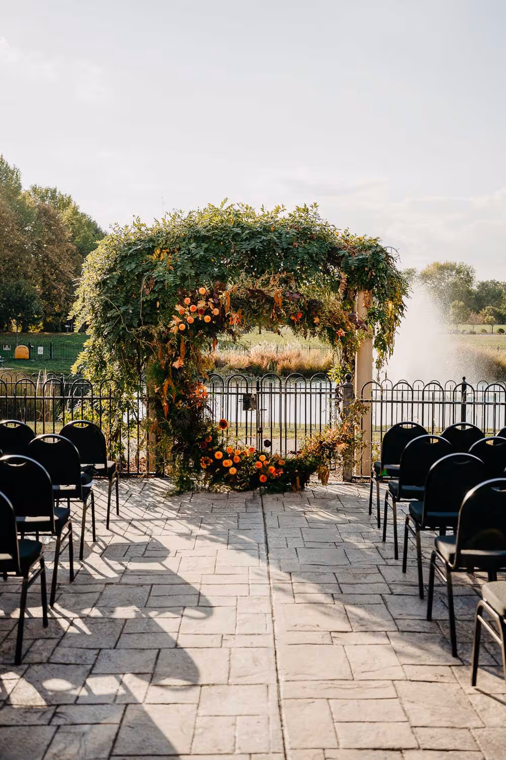Outdoor wedding ceremony setup with black chairs facing a circular floral arch decorated with green foliage and orange flowers near a pond with a fountain.