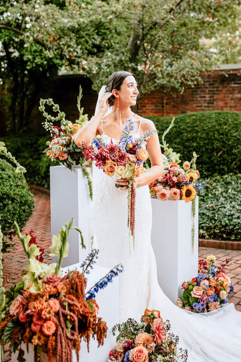 Bride in white lace dress holding a colorful bouquet, standing outdoors among various flower arrangements on white pedestals.
