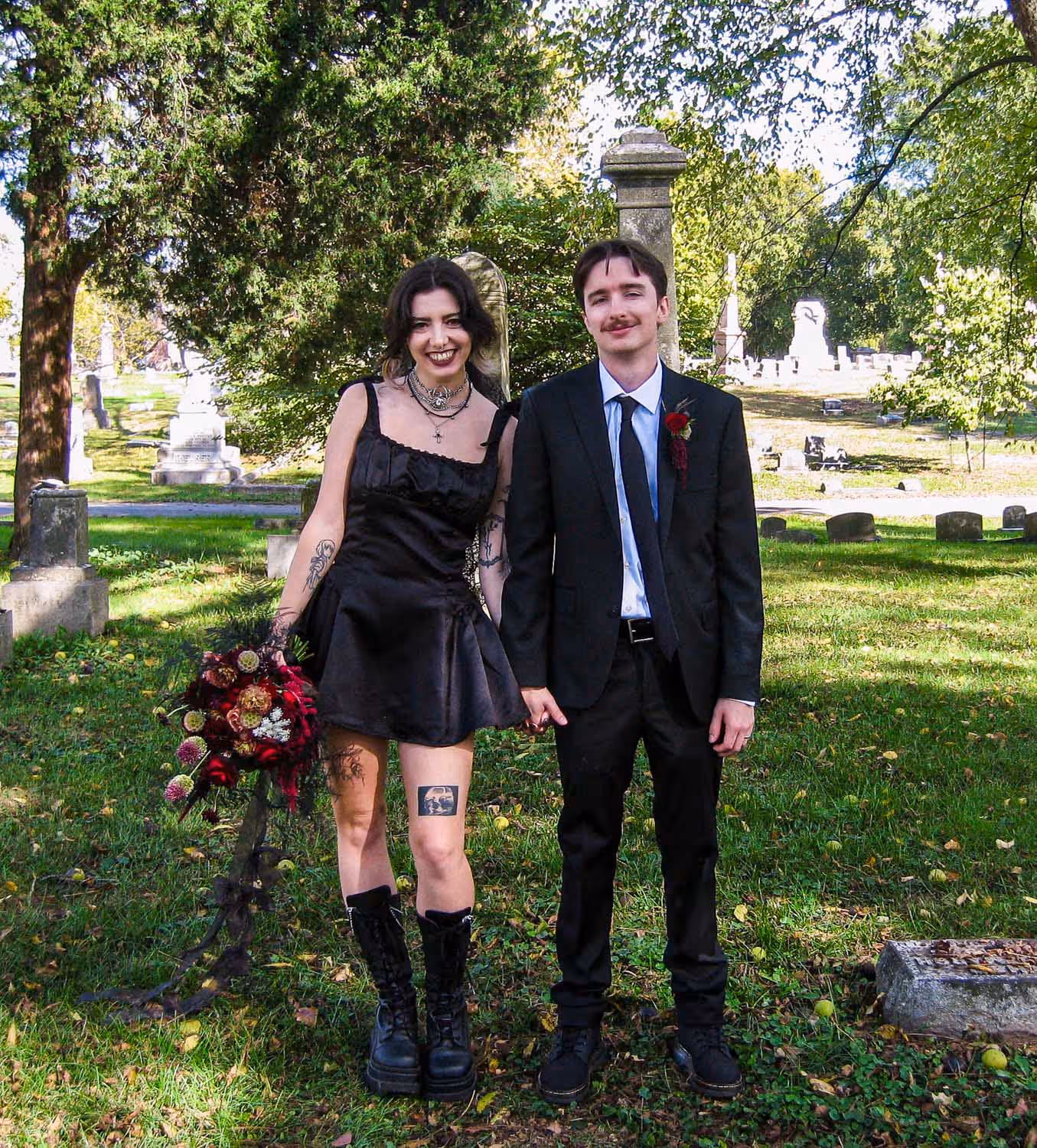 A couple holding hands in a cemetery; the woman wears a black dress and boots holding a bouquet, the man wears a black suit with a red boutonniere.