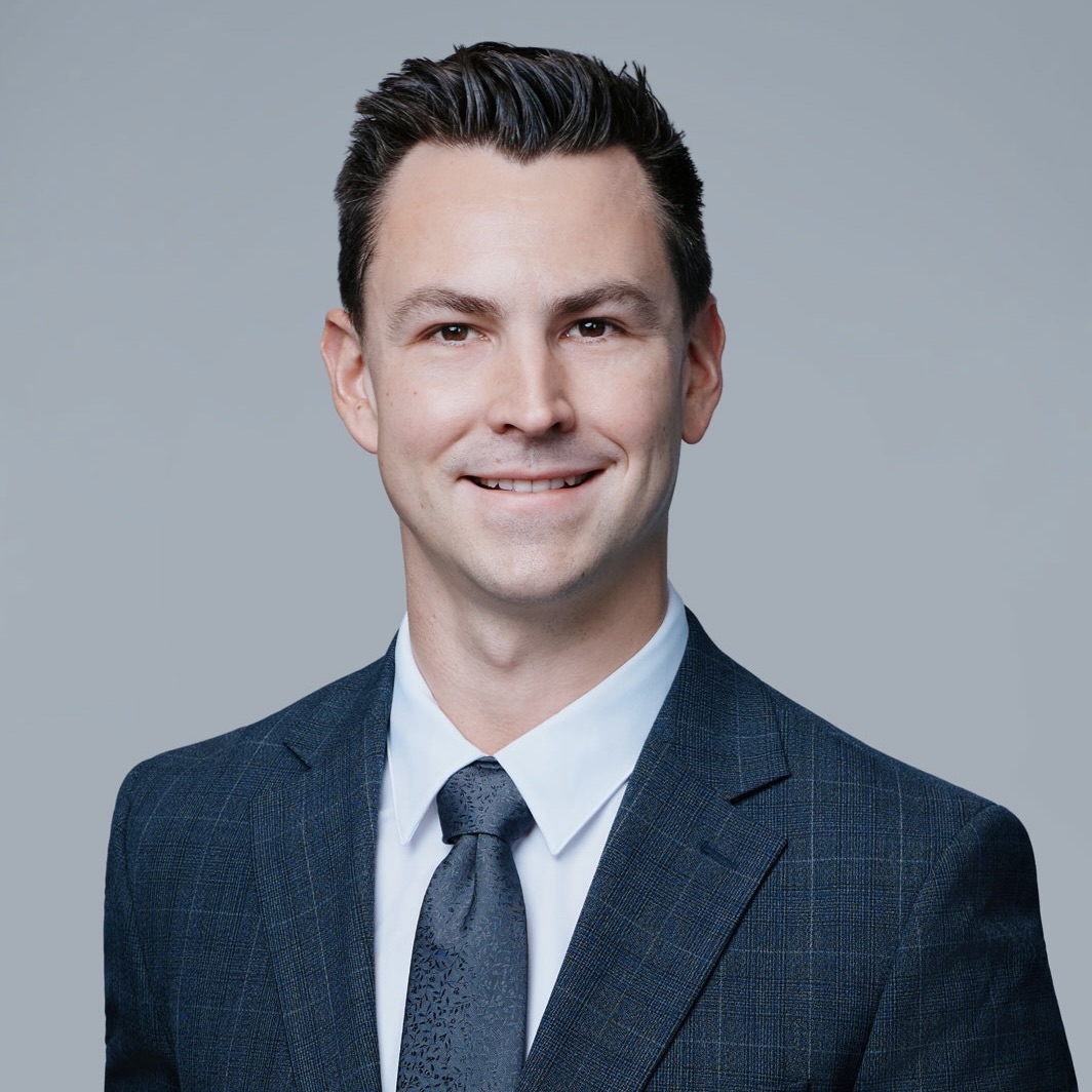 Smiling man wearing a dark blue suit, white shirt, and patterned tie against a plain gray background.