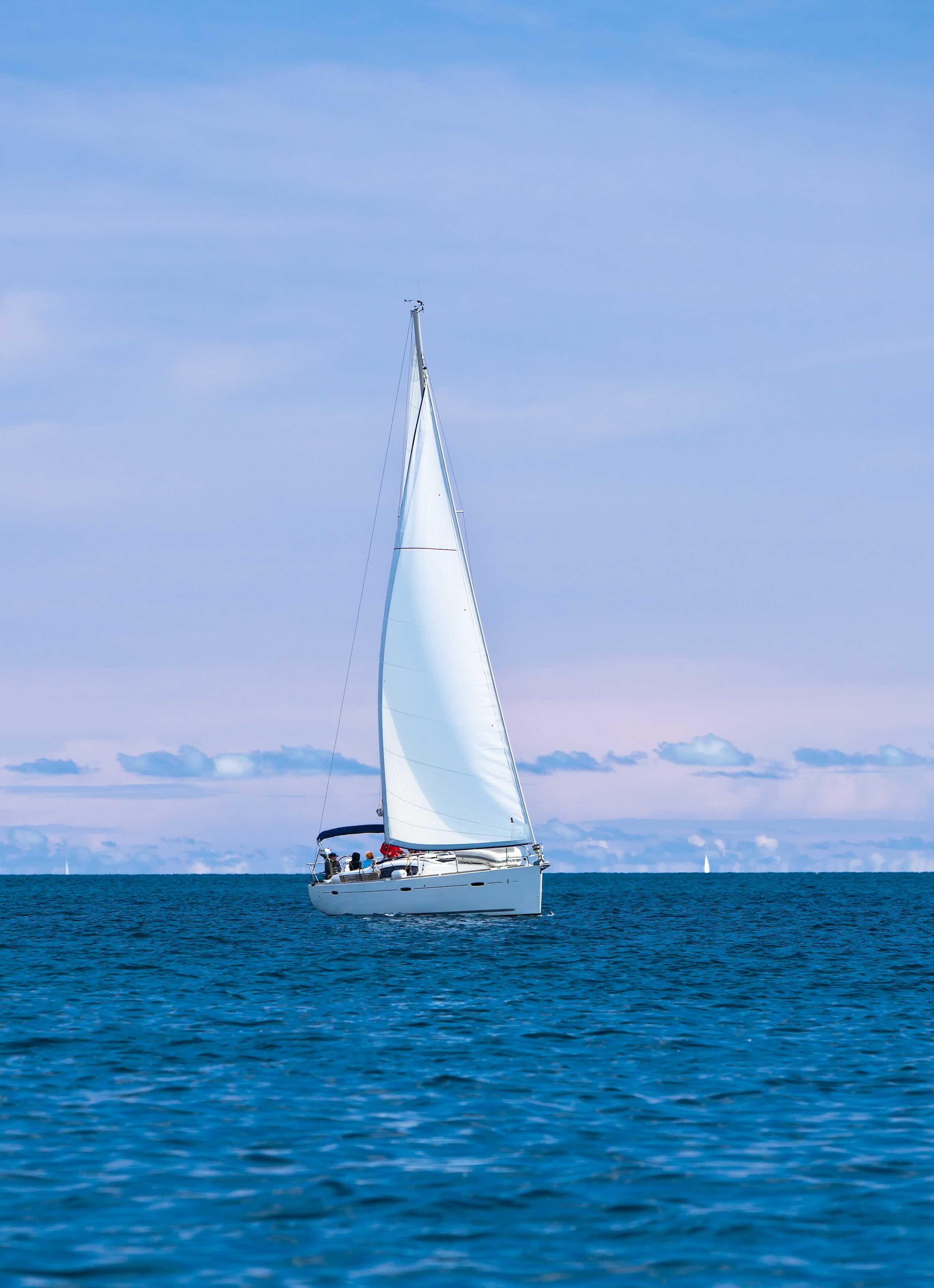 White sailboat on calm blue ocean under cloudy sky with people on board.