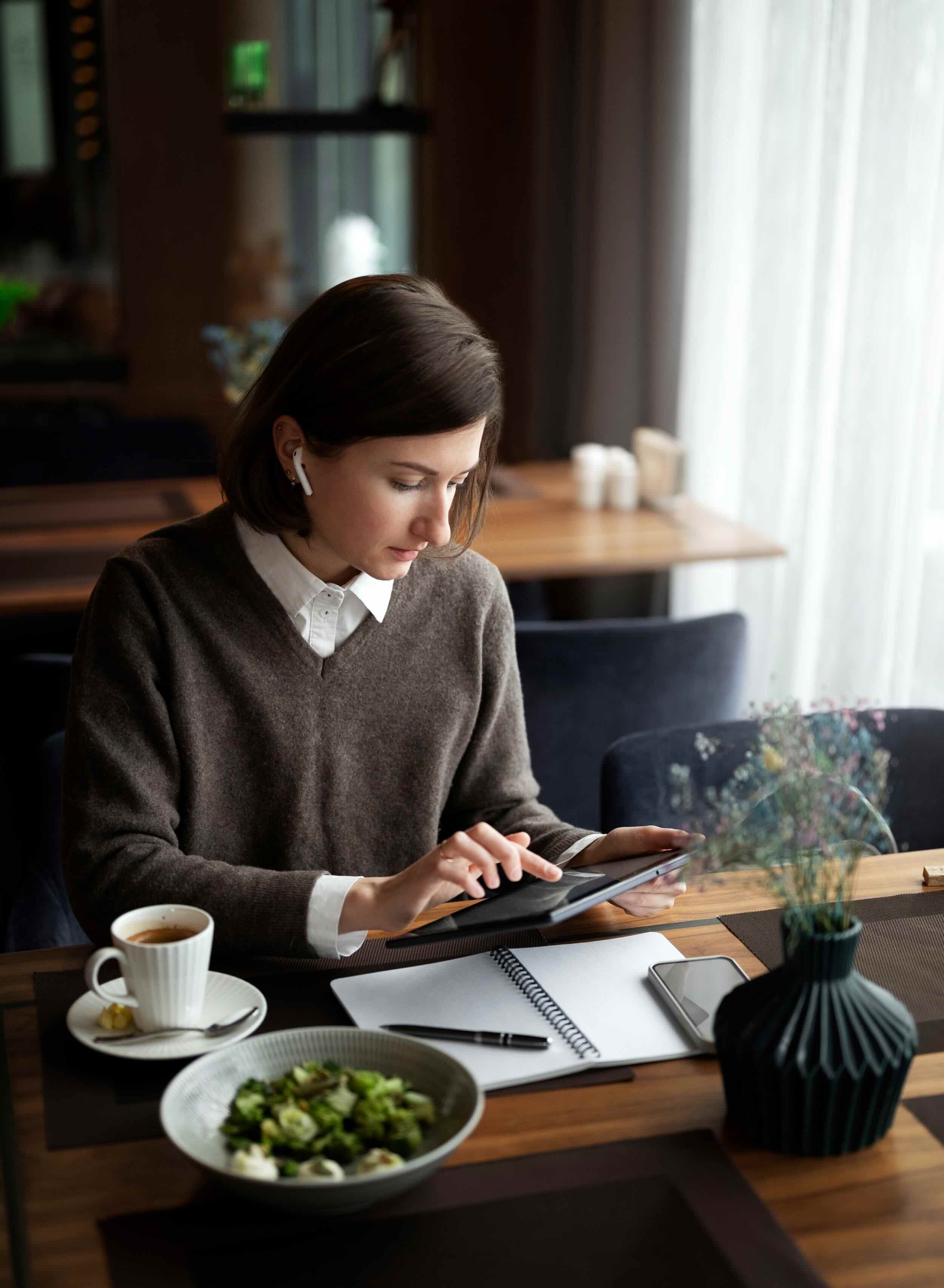 Woman wearing earbuds using a tablet at a table with notebook, phone, coffee, and salad bowl.