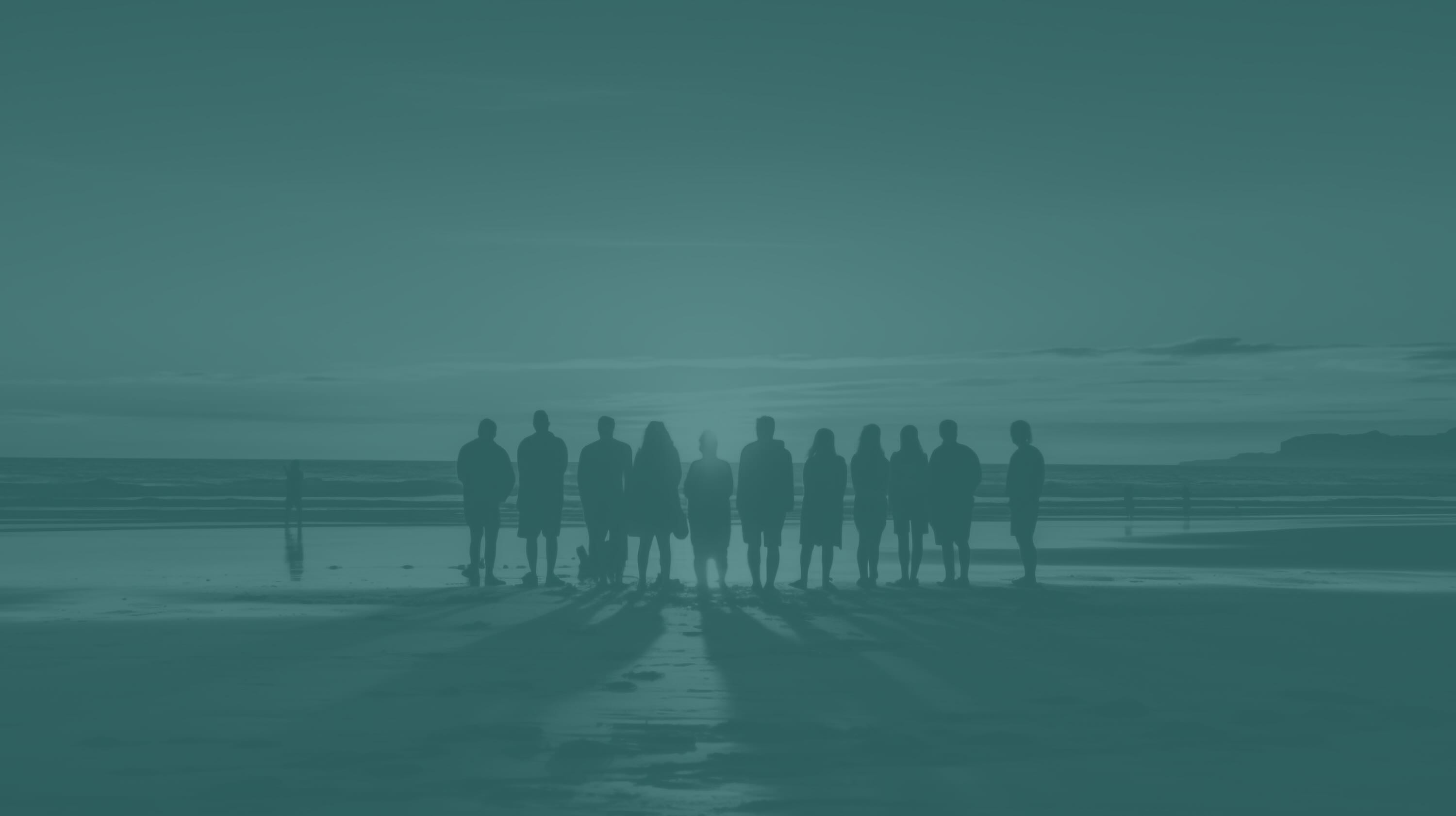 Silhouettes of eleven people standing on a beach at sunset with long shadows on wet sand.