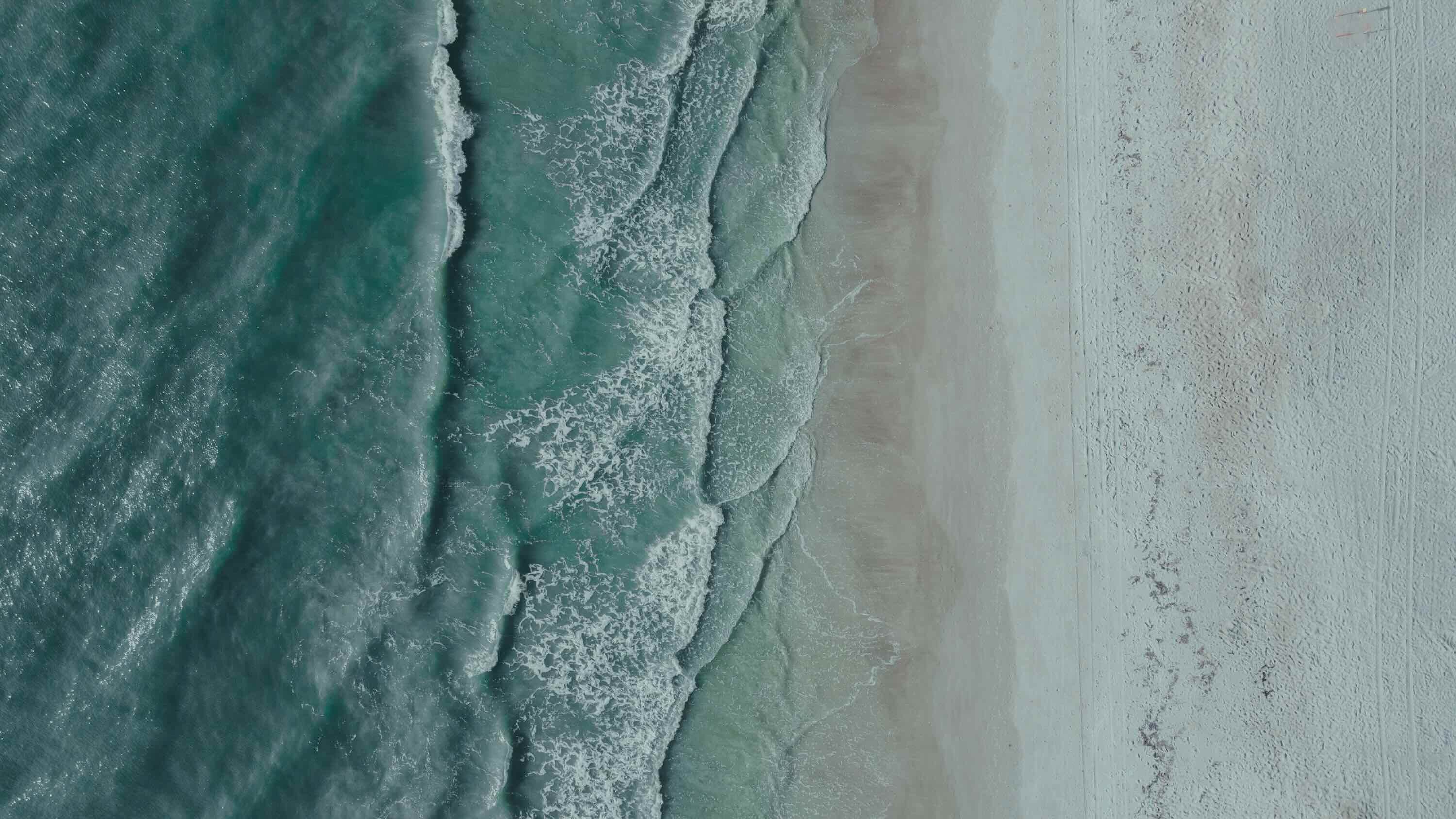 Aerial view of ocean waves gently rolling onto a sandy beach with visible tire and foot tracks.