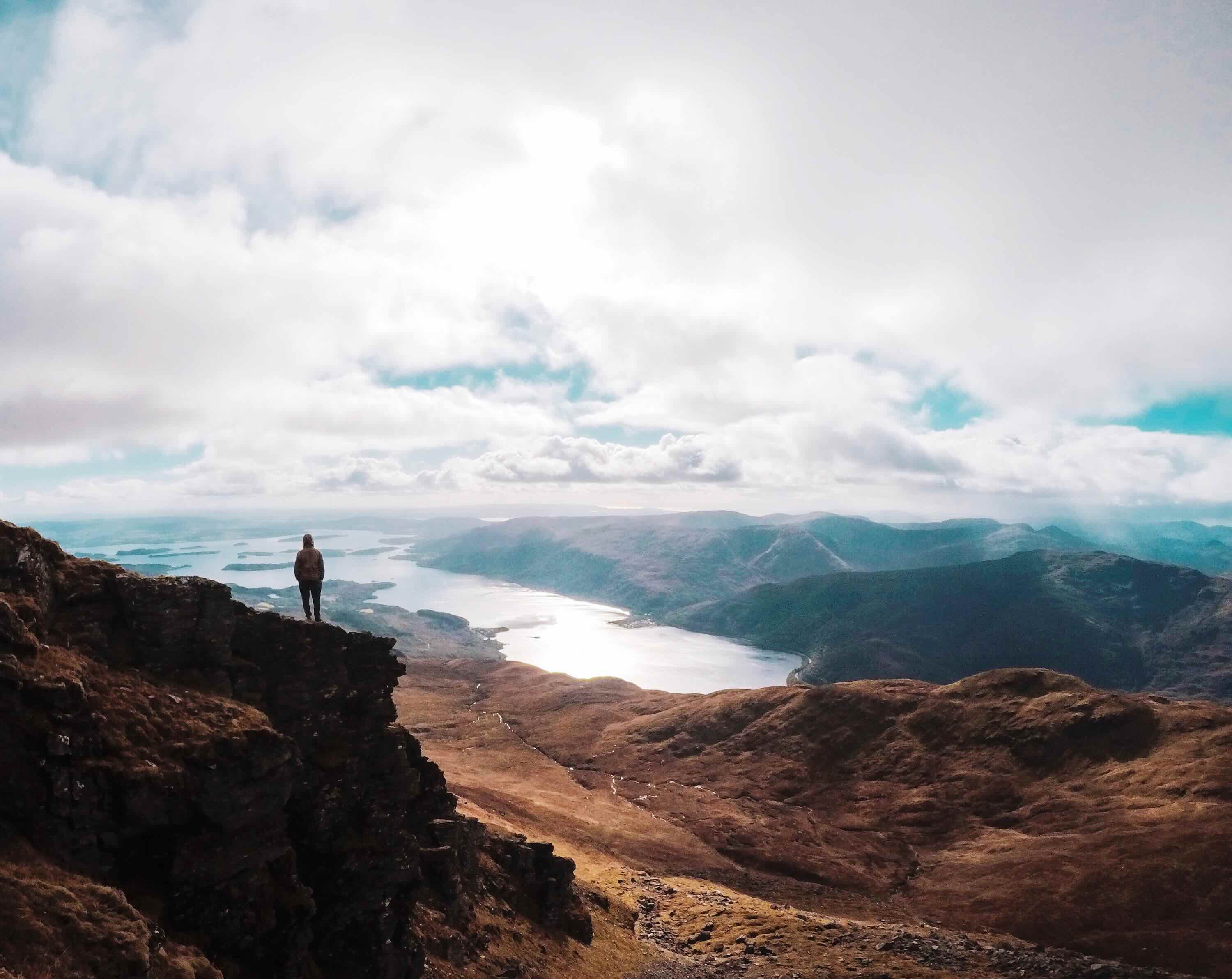 Person standing on rocky cliff overlooking lake and mountain landscape under cloudy sky.