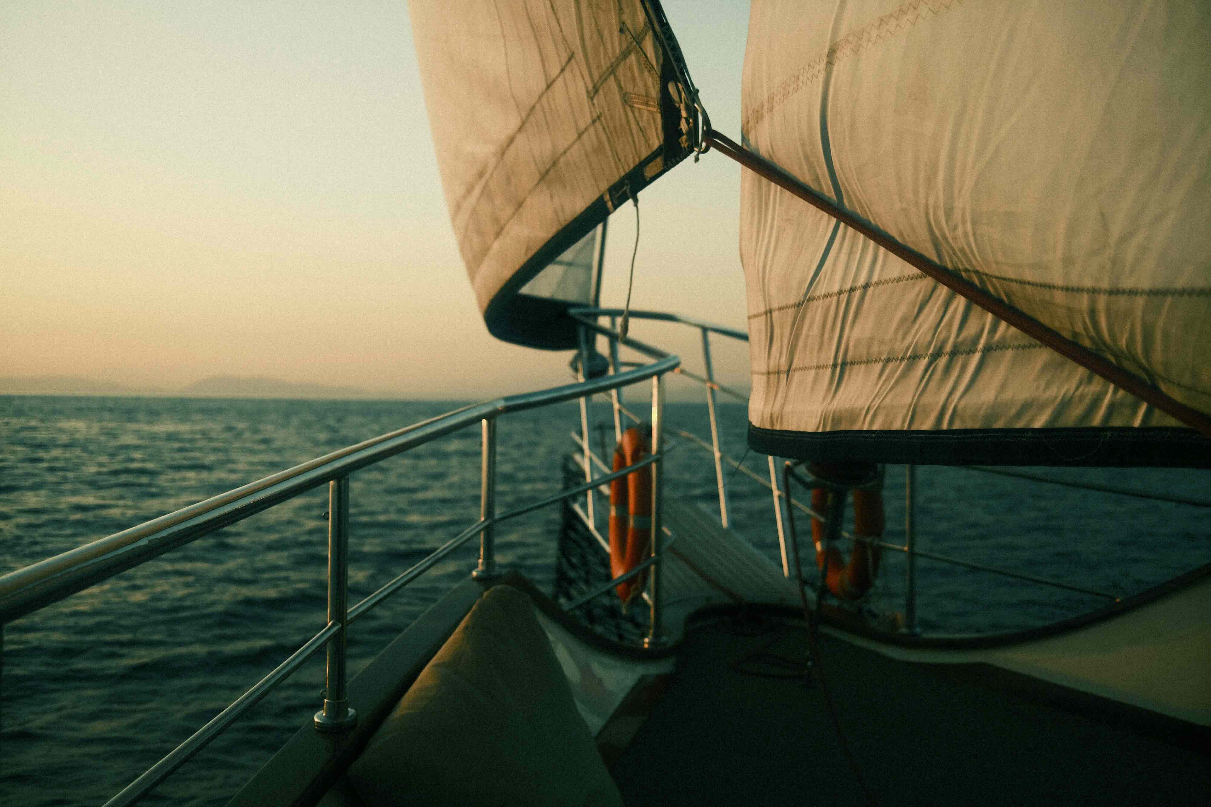 View along a sailboat railing with sails up on calm water at sunset.