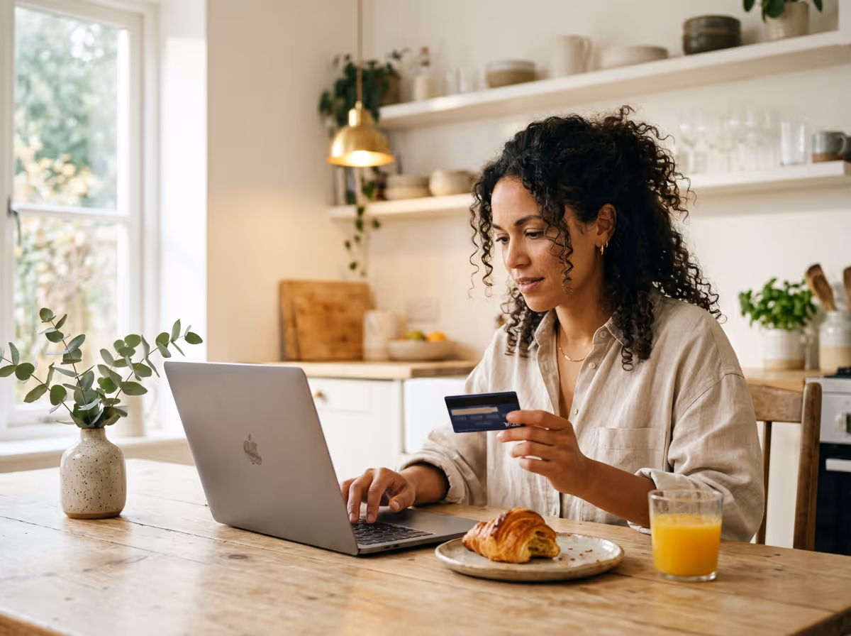 Woman sitting at kitchen table using laptop and holding a credit card with croissant and orange juice on table.