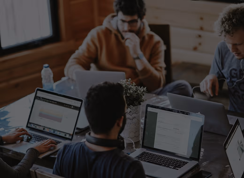 Group of people working on laptops around a table with a small potted plant in the center.
