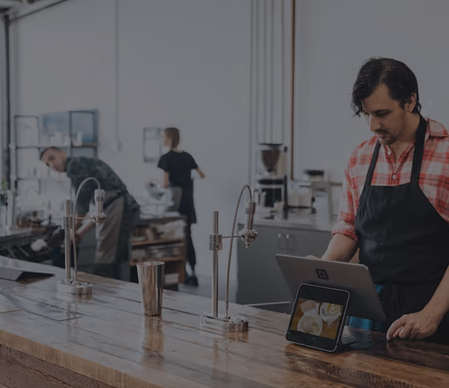 Barista wearing apron working on a tablet behind a wooden counter in a coffee shop.