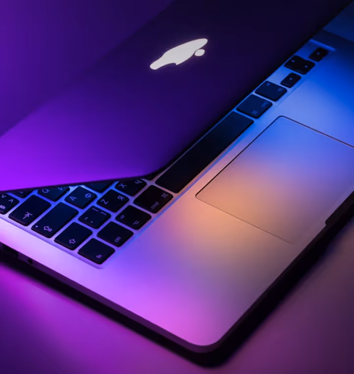 Partially open Apple laptop illuminated by purple and orange lighting, showing keyboard and trackpad.