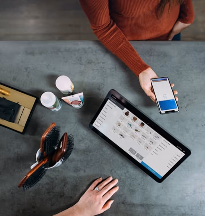 Overhead view of two people using a tablet and phone with a hairbrush and beauty products on table.